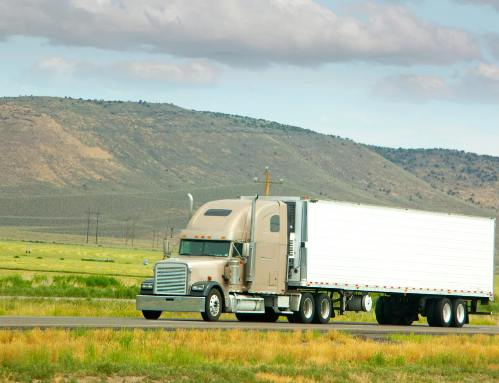 Freight-Hauler-tan-semi-truck-driving-down-highway-with-meadowy-mountains-behind-it