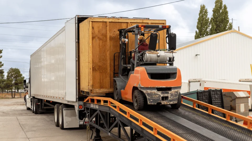 Moving Freight man operating orange forklift unloading huge wooden crate out of white semi trailer